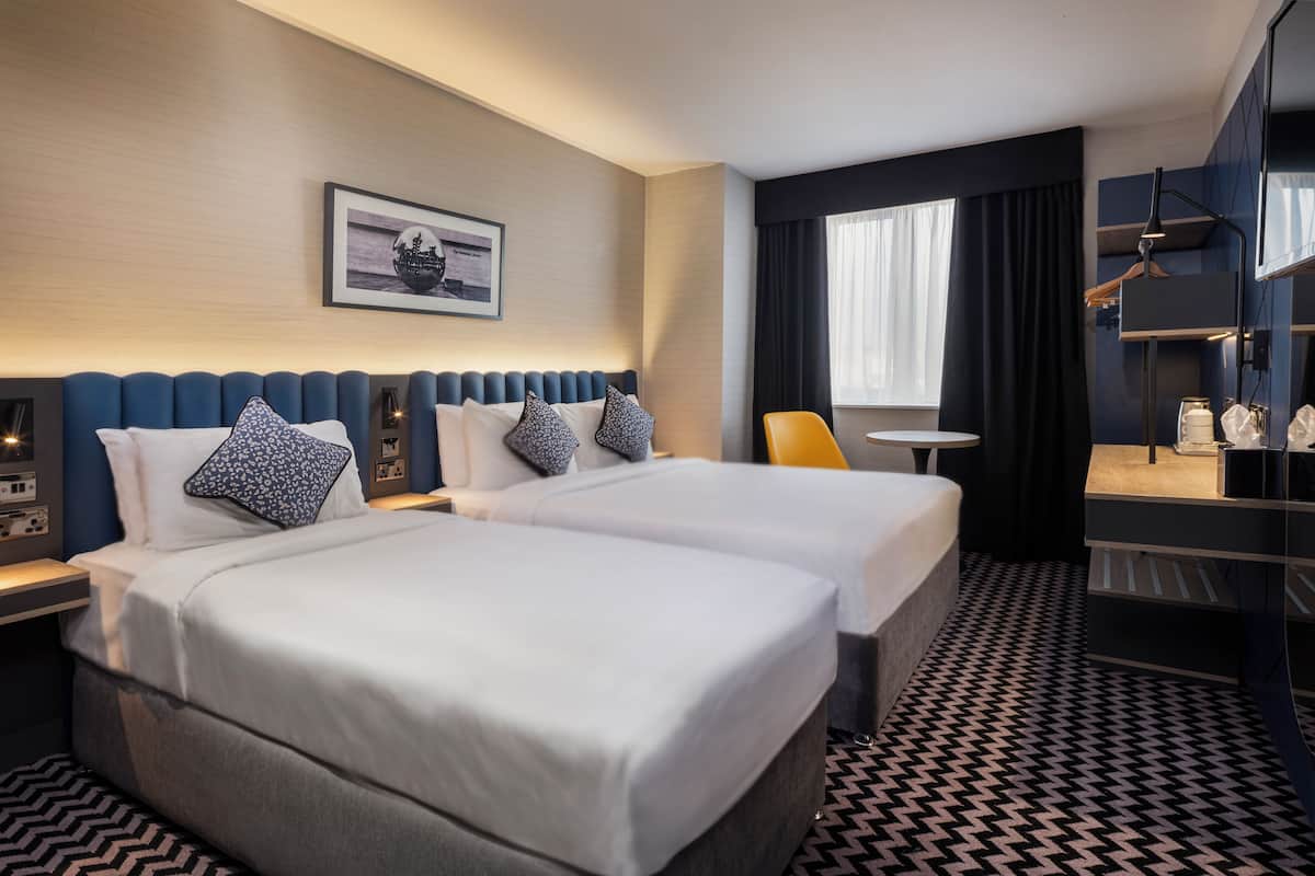 A hotel room with two beds, a desk, a chair, a small table, and a TV.