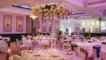 A banquet hall with round tables set for a formal event, decorated with floral arrangements and chandeliers.