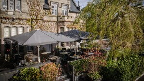 Un caf é al aire libre con mesas y sombrillas, frente a un edificio histórico de paredes de piedra y hiedra.
