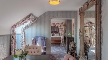 A room with a large mirror, a tufted chair, a bed with floral curtains, and a vase with flowers on a table.