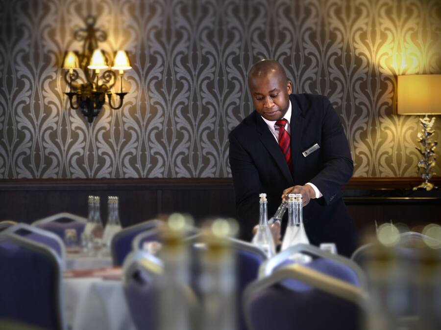 A man in a suit preparing a table in a room with patterned wallpaper and chandeliers.