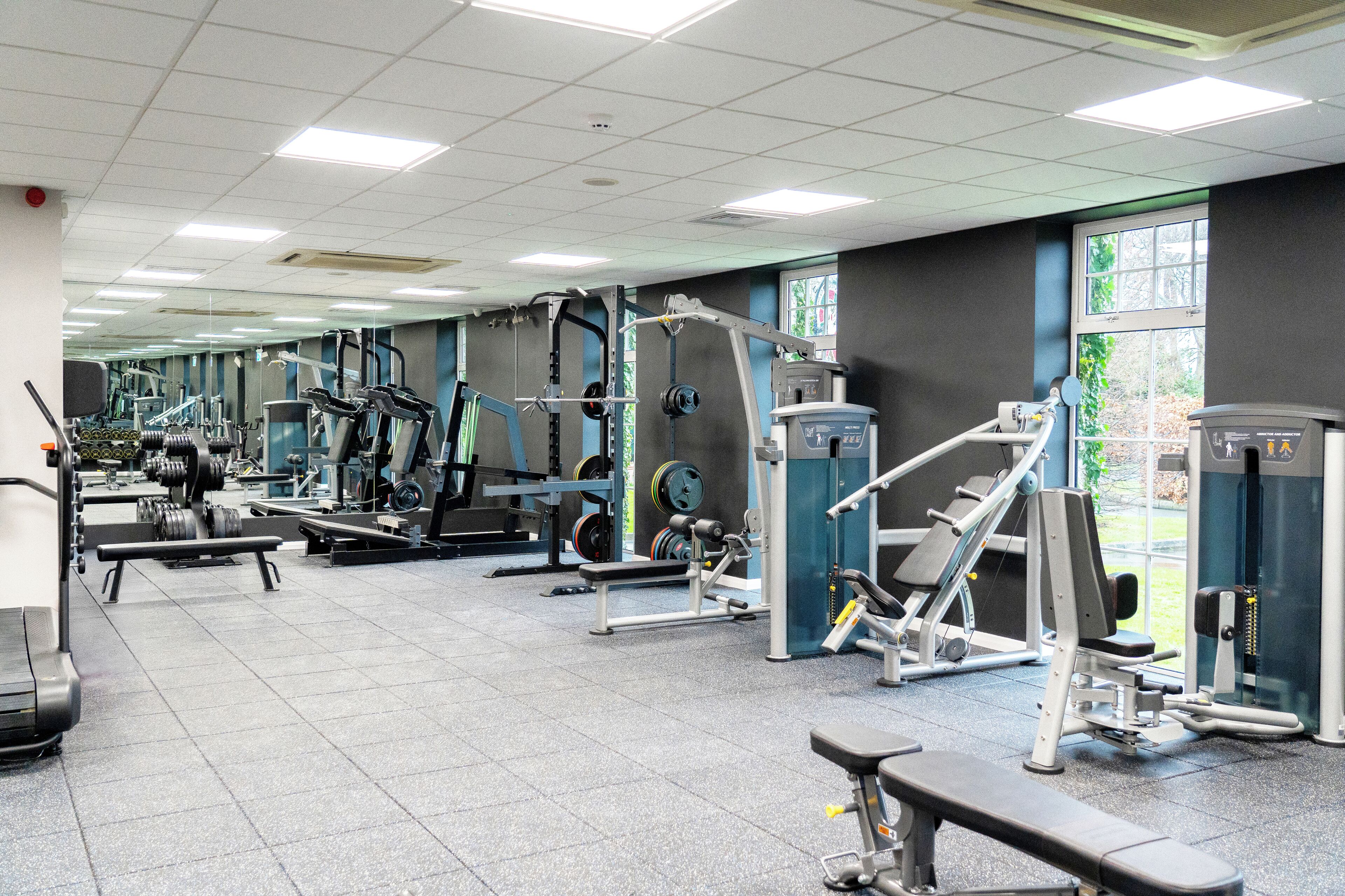 Un gimnasio con diversas máquinas de ejercicio, una banca y un ventanal que proporciona luz natural.
