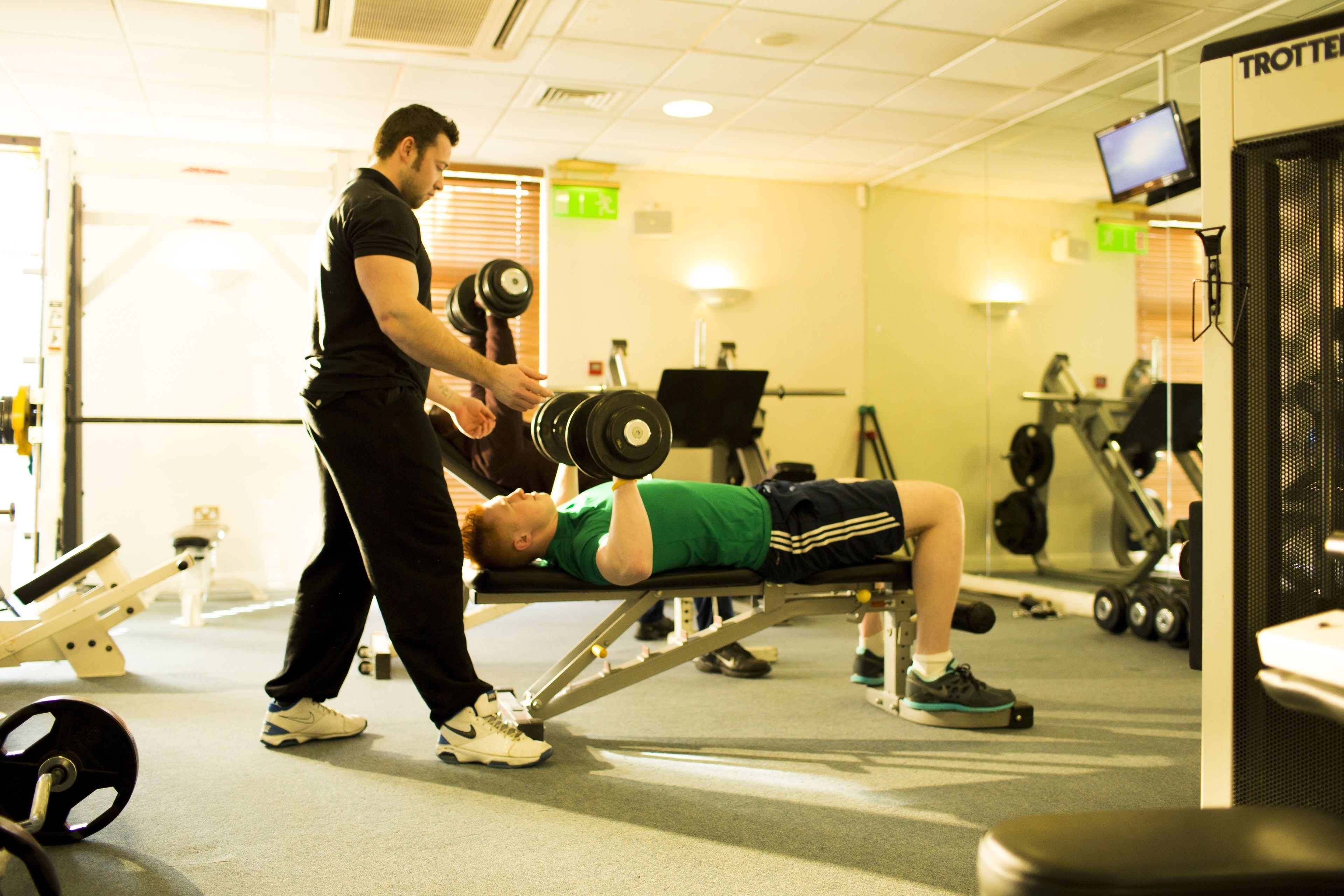 A gym with exercise equipment, a person lying on a bench, and another person assisting.