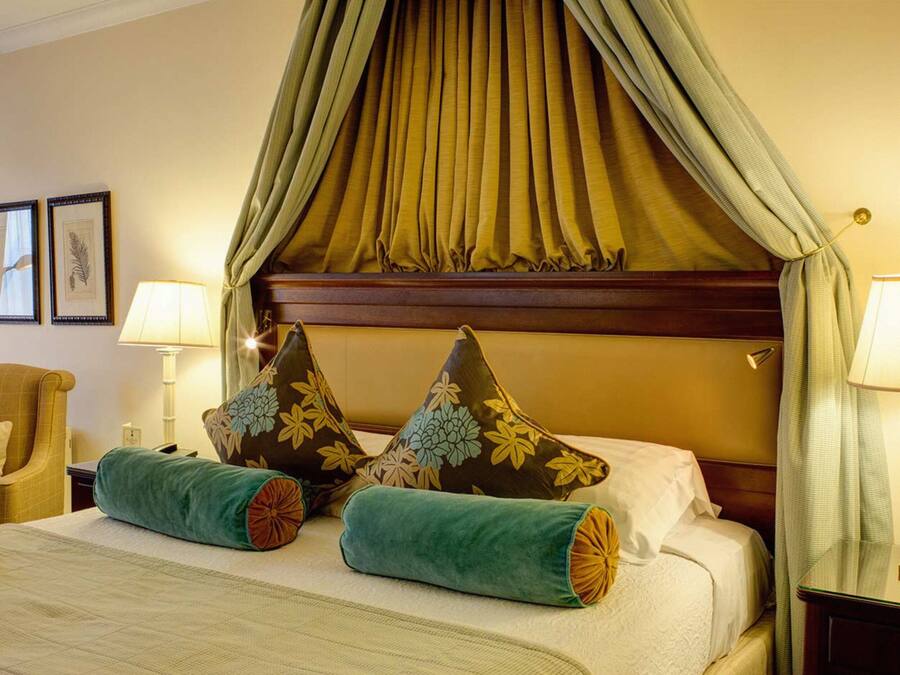 A hotel room with a canopy bed, two pillows with floral designs, a green throw pillow, a chair, and two bedside lamps.