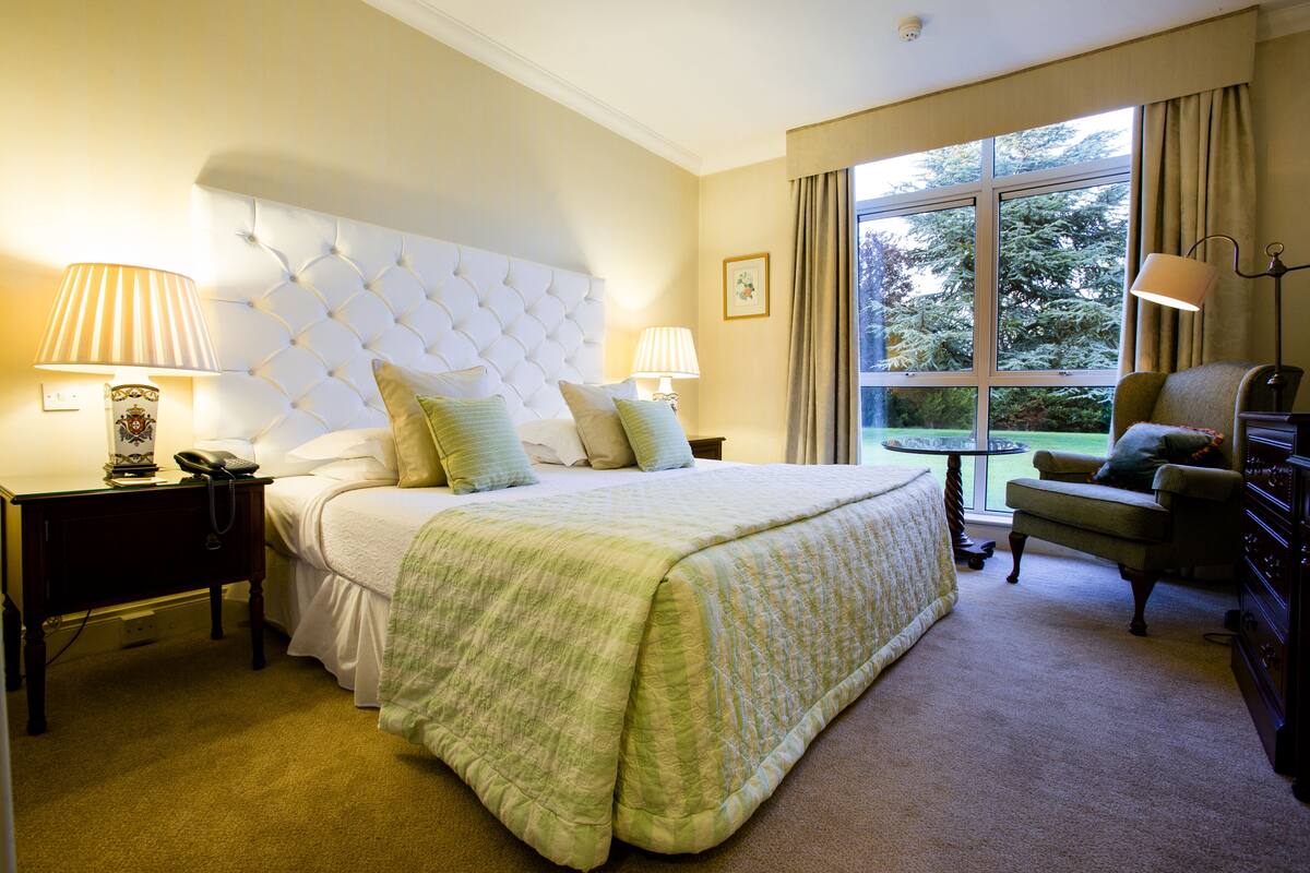 A hotel room with a large bed, two bedside tables with lamps, a chair, and a window with curtains.