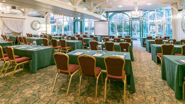 Un salón de conferencias con varias mesas y sillas, ventanas amplias y una araña.
