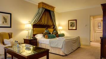 A hotel room with a four-poster bed, a sitting area with a chair and a table, and a door leading to another room.