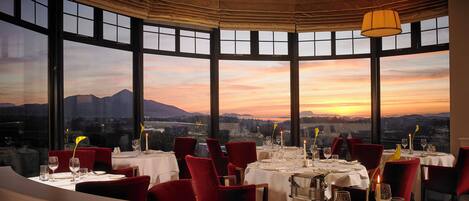 A restaurant with a table set for a meal, large windows, and a view of the sunset.
