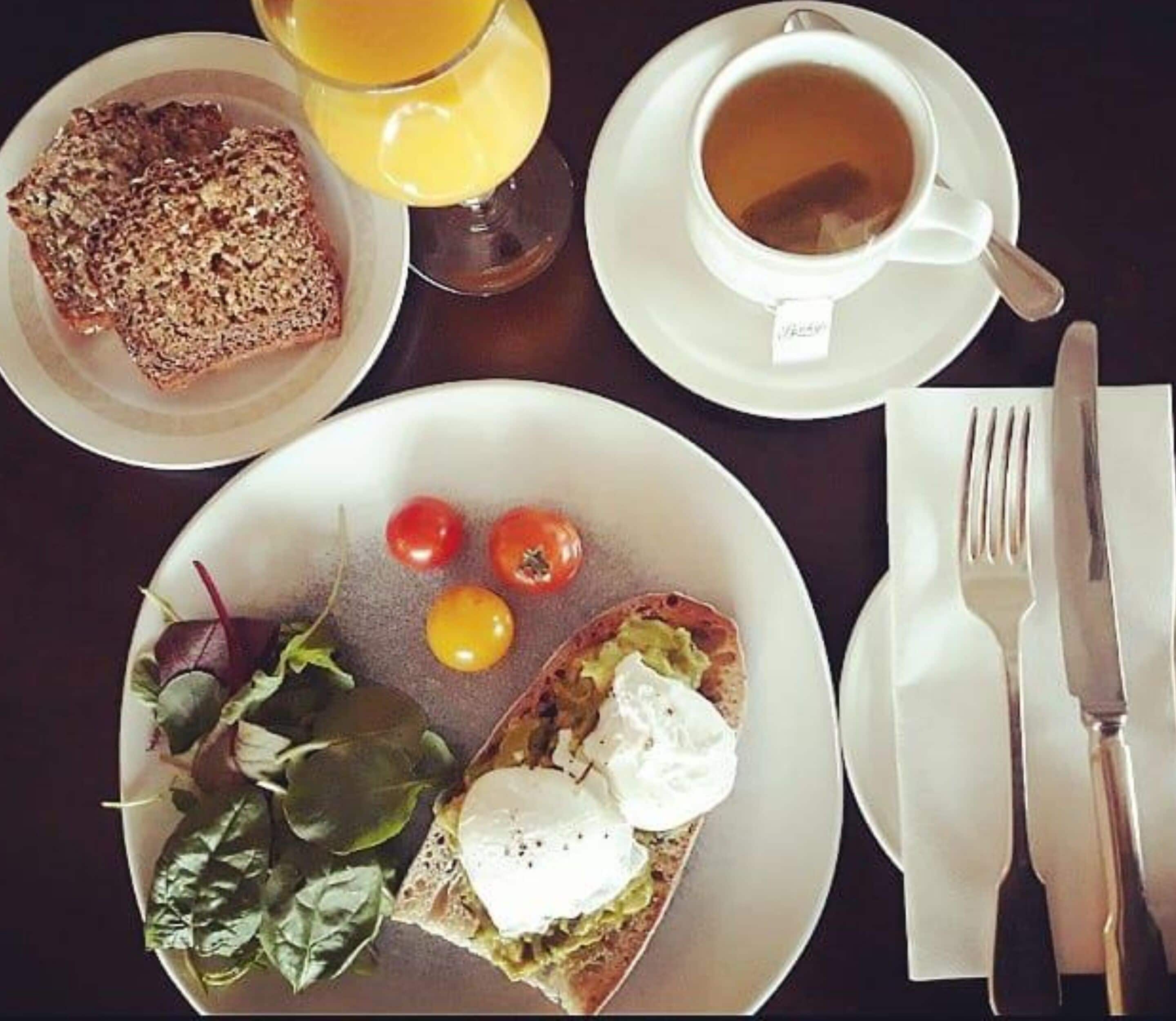 A breakfast setting with toast, tomatoes, spinach, and a poached egg on a plate, a cup of tea, and a glass of orange juice on a table.
