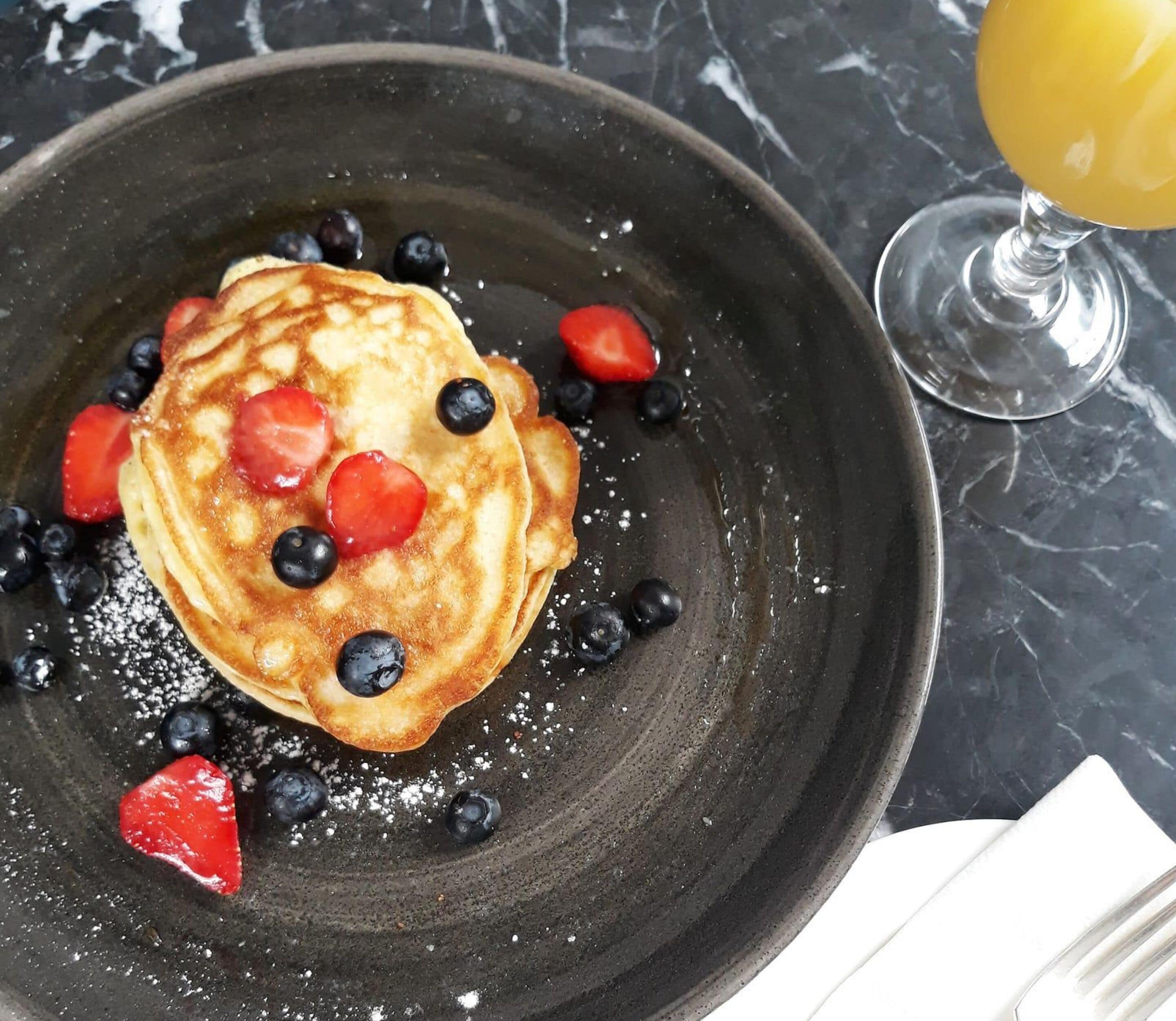 A plate of pancakes with blueberries and strawberries, a glass of orange juice, and a marble countertop.
