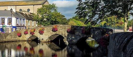 A stone bridge over a calm river with flower pots on the sides, a building with a blue facade, and trees in the background.