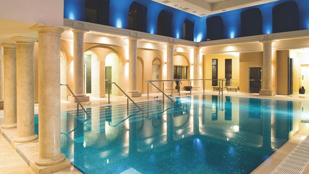 Una amplia piscina interior con agua cristalina y azul, rodeada de columnas y arcos, e iluminada con luz cenital.