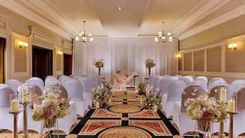 A formal event space with rows of white chairs, a central aisle with floral arrangements, and chandeliers.