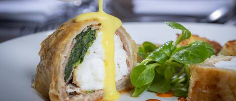 A dish with a creamy sauce being poured over it, accompanied by greens and a piece of meat.