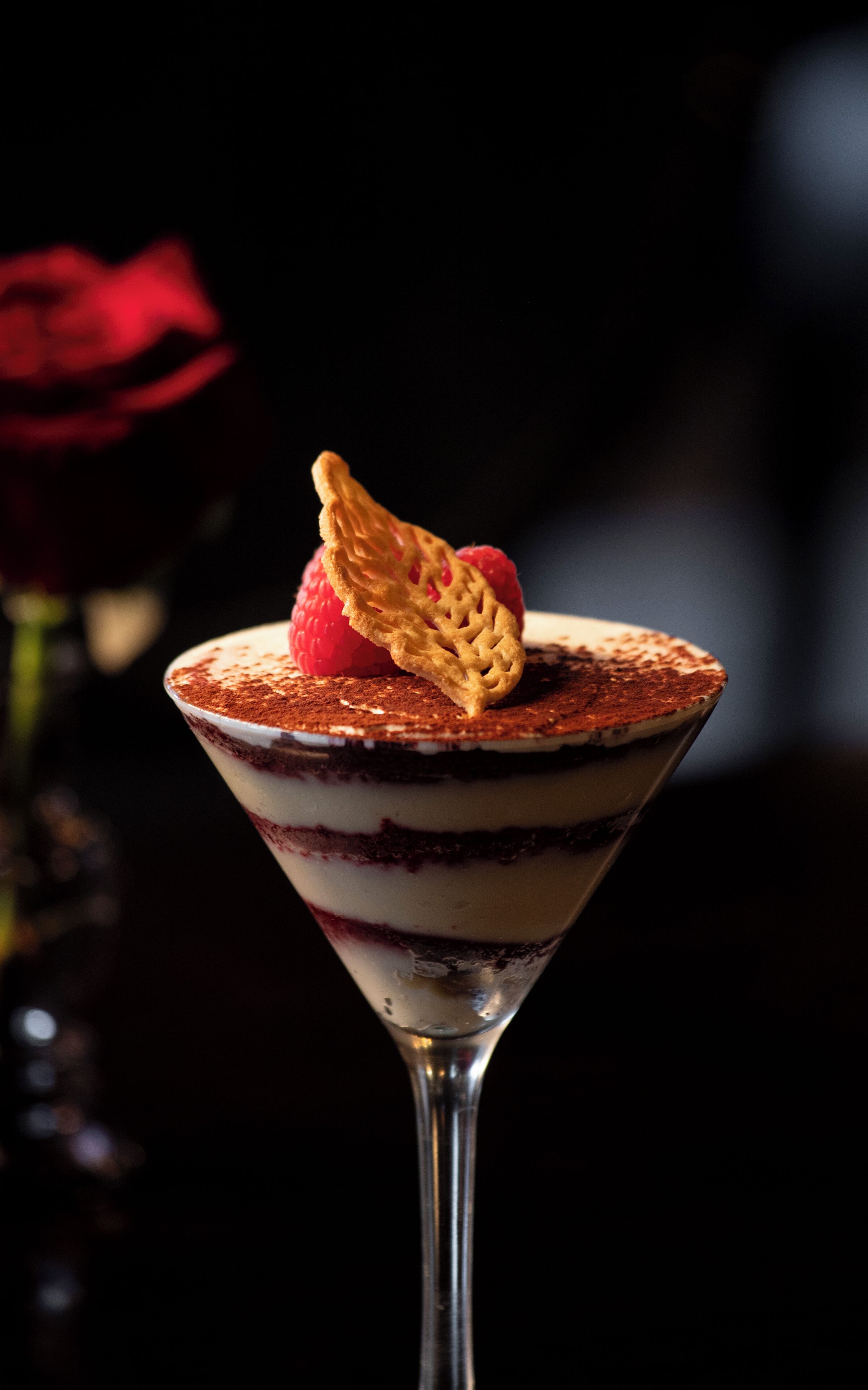 A dessert in a martini glass with layers of cream and chocolate, a raspberry, and a wafer.