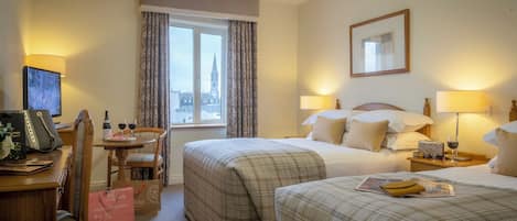 A hotel room with two beds, a desk with a chair, a window with curtains, a framed picture on the wall, and a lamp on the bedside table.