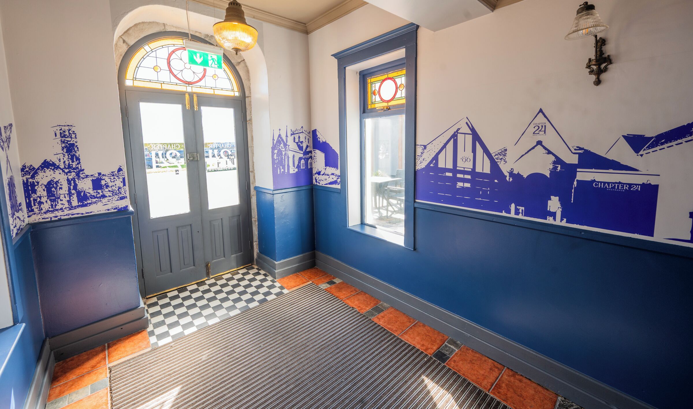 Un corridoio con pavimento a scacchi, una porta con vetrata colorata e pareti decorate con affreschi blu.