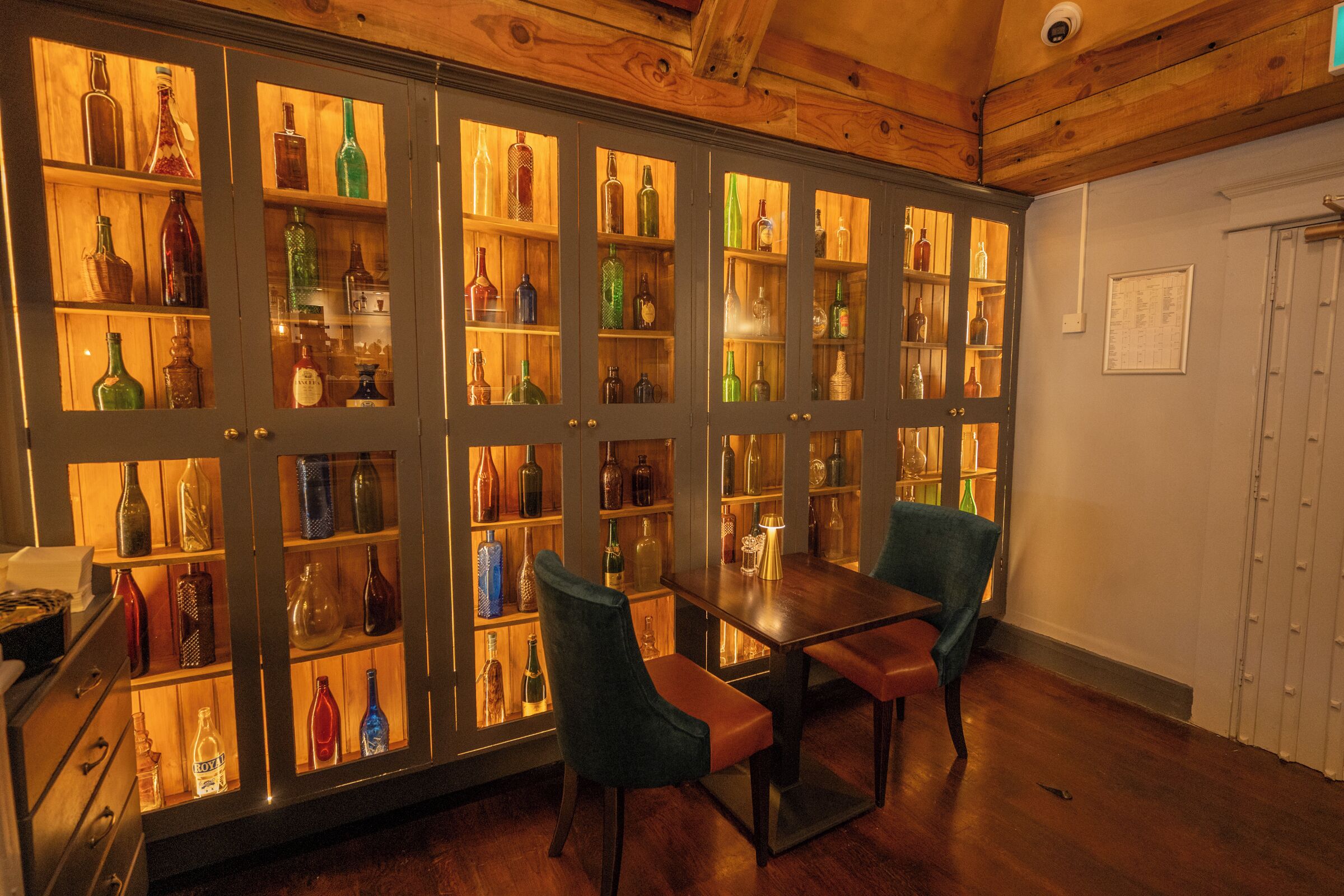 A room with a wooden ceiling, a table with chairs, and a cabinet with glass doors displaying various bottles.