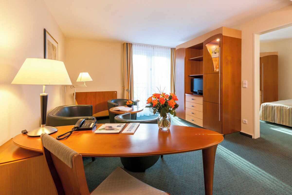 A hotel room with a wooden desk, two chairs, a telephone, a vase with flowers, a television, and a bed.