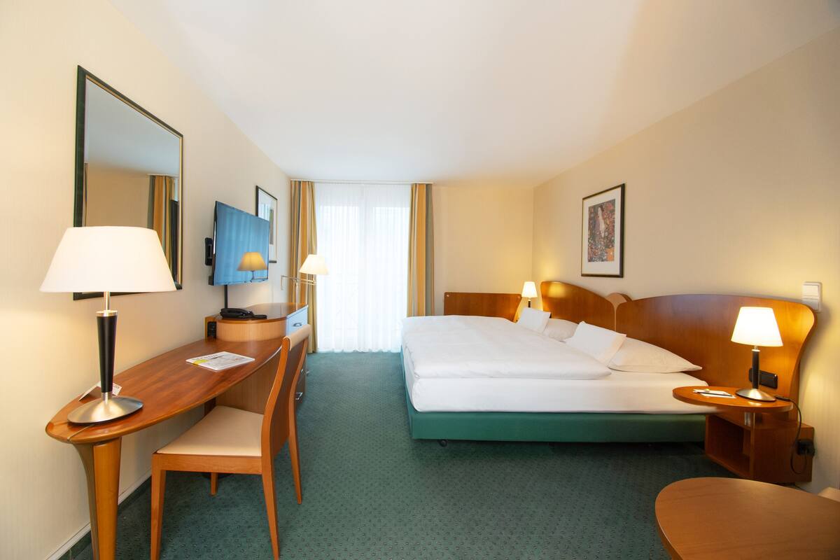 Hotelkamer met een bed, een bureau met stoel, bureaulamp, nachtkastje met lamp, en een grote spiegel aan de muur.