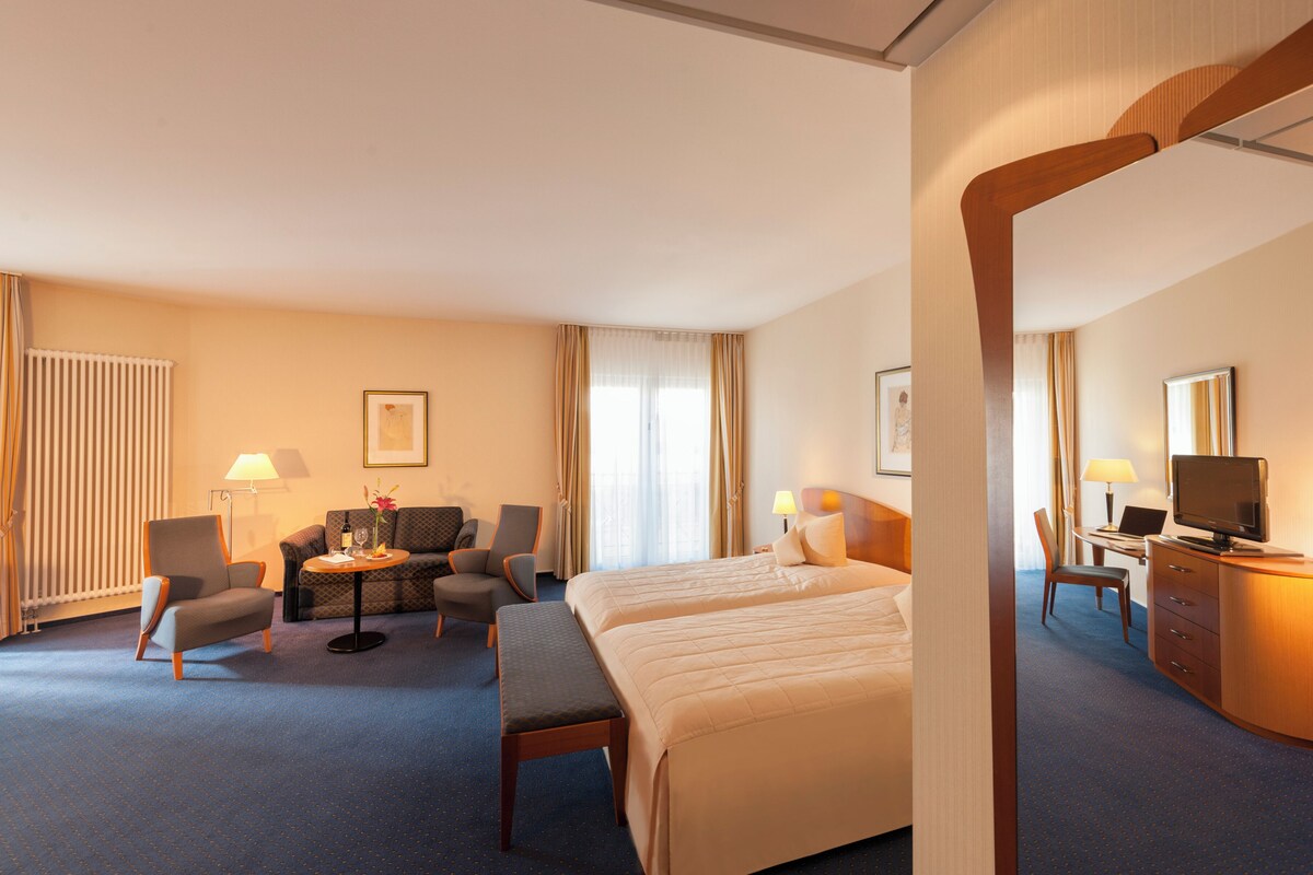 A hotel room with a bed, two armchairs, a small table, a desk with a computer, a lamp, and a window with curtains.