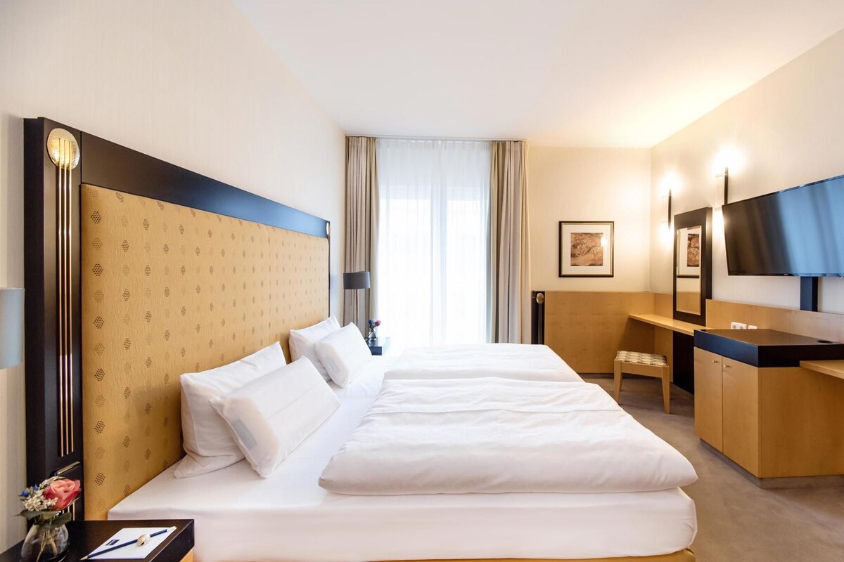 Ein Hotelzimmer mit einem großen Bett, einem Schreibtisch, einem Sessel, einem Fernseher und einem Fenster mit Vorhängen.