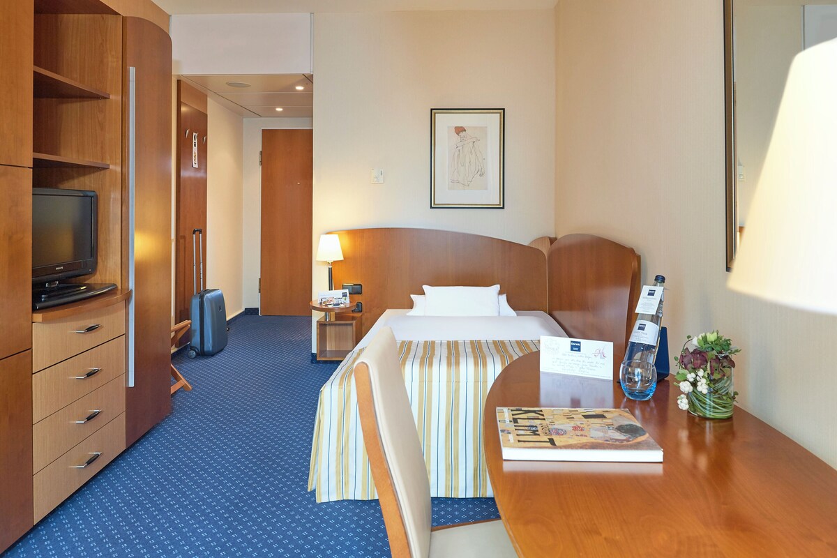 A hotel room with a bed, a desk with a lamp, a television, a suitcase, and a framed picture on the wall.
