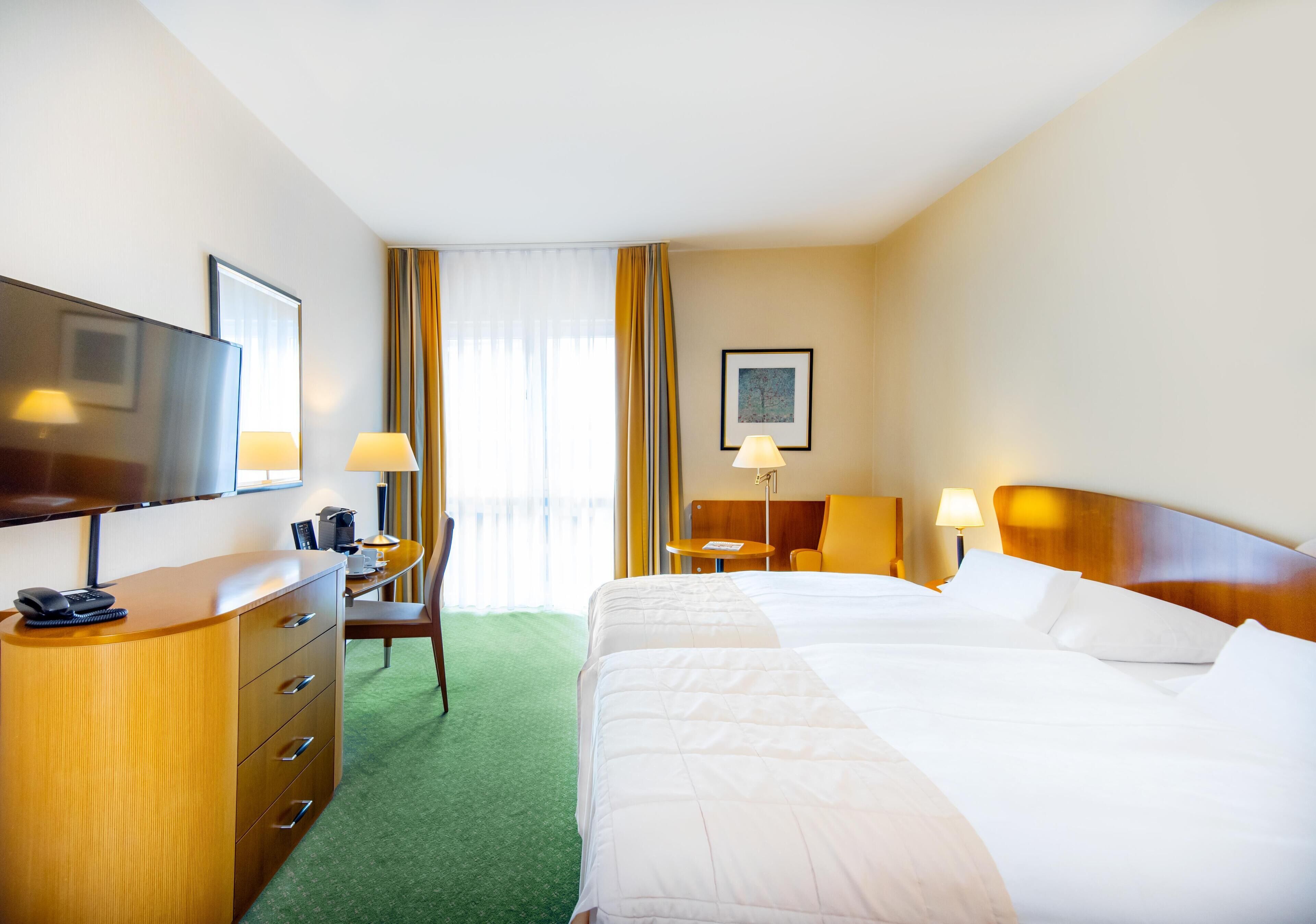 A hotel room with a bed, a desk with a chair, a television, and a window with curtains.