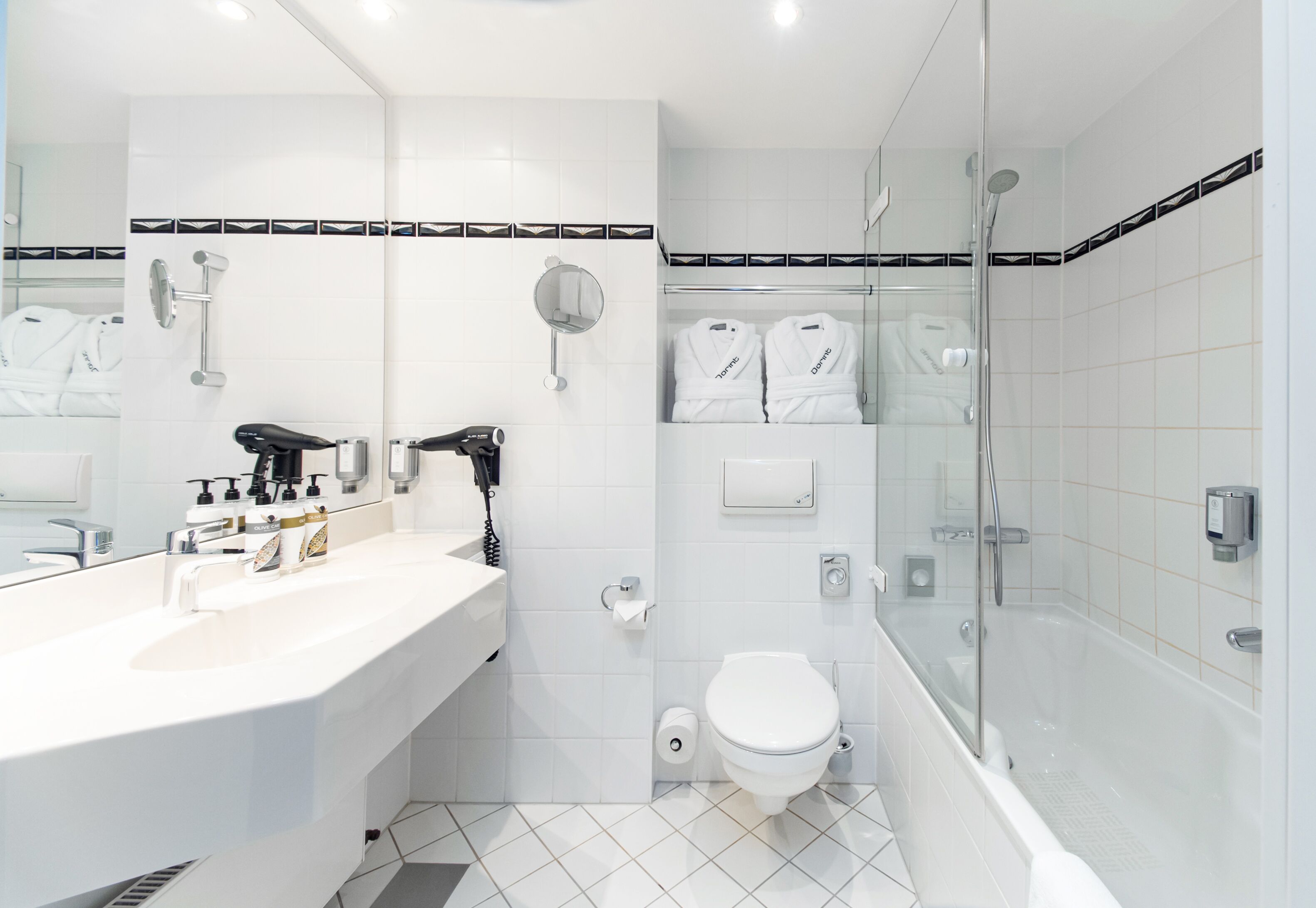 Un baño moderno con lavamanos, espejo y váter blancos. Hay dos albornoces blancos colgados en la pared, un box de ducha de cristal y una bañera blanca.