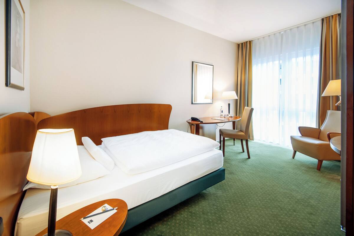 Een hotelkamer met een bed, nachtkastje, lamp, stoel, bureau en een groot raam met gordijnen.