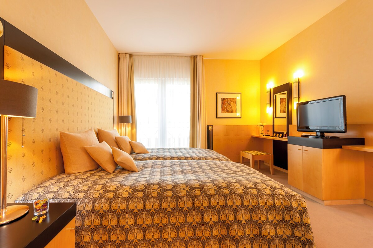 A hotel room with a bed, bedside table, lamp, TV, and a small stool.