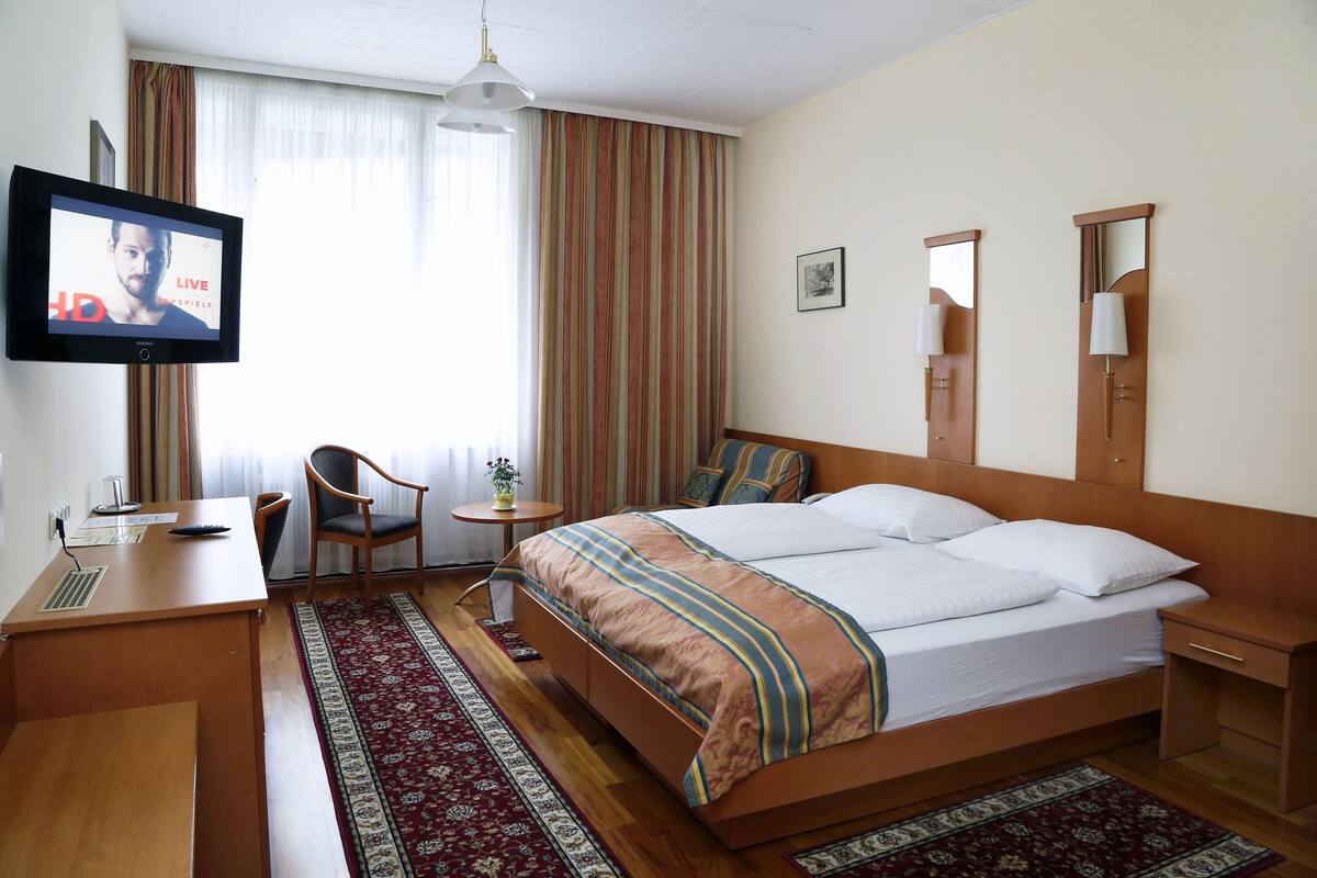 A hotel room with a bed, a desk, a chair, a TV, and a window with curtains.