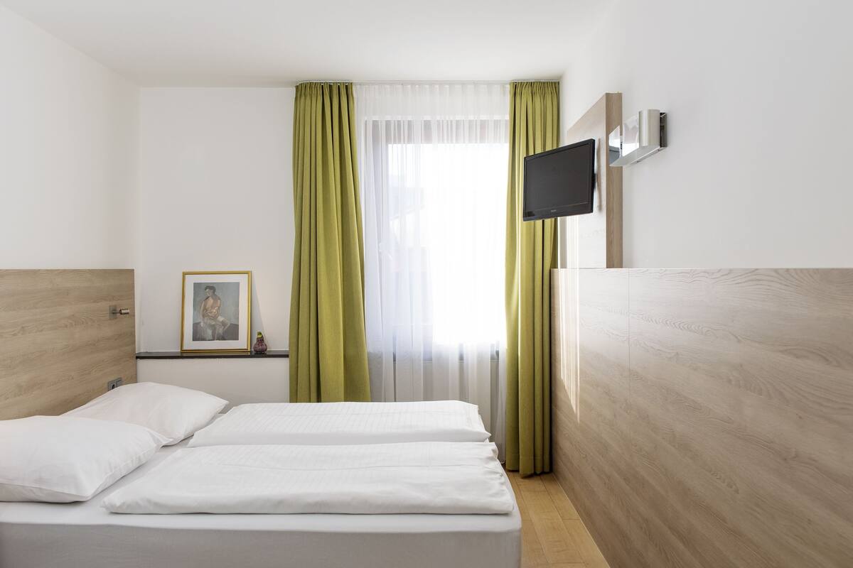 A hotel room with two beds, a TV mounted on the wall, a framed picture on the wall, and a window with green curtains.