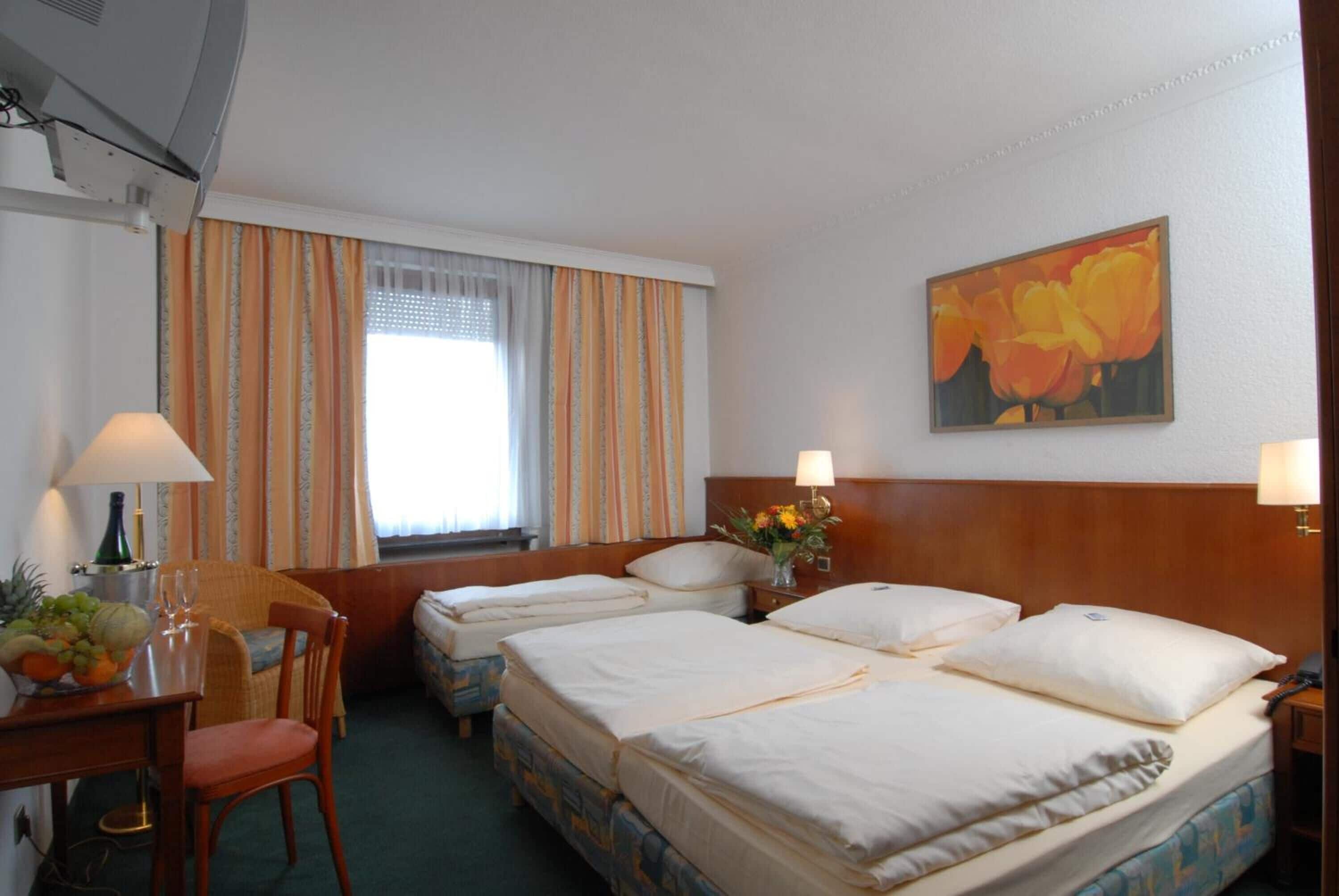 Una habitación de hotel con dos camas, un escritorio con lámpara, una silla, una mesita con fruta y un florero, y un cuadro en la pared.