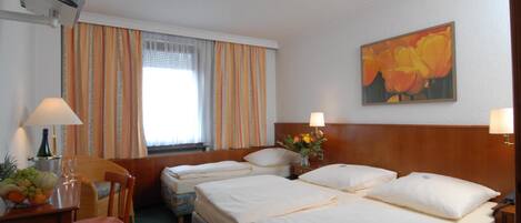 Una habitación de hotel con dos camas, un escritorio con lámpara, una silla, una mesita con fruta y un florero, y un cuadro en la pared.