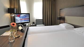 Une chambre d’hôtel avec deux lits, une télévision, un bureau avec une lampe, un vase avec une fleur et des rideaux.