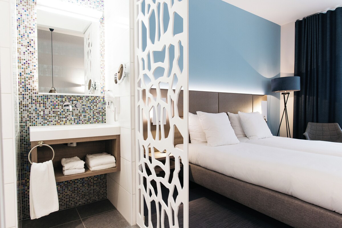 A modern hotel room with a large bed, a bathroom with a mosaic tile backsplash, and a decorative white partition.