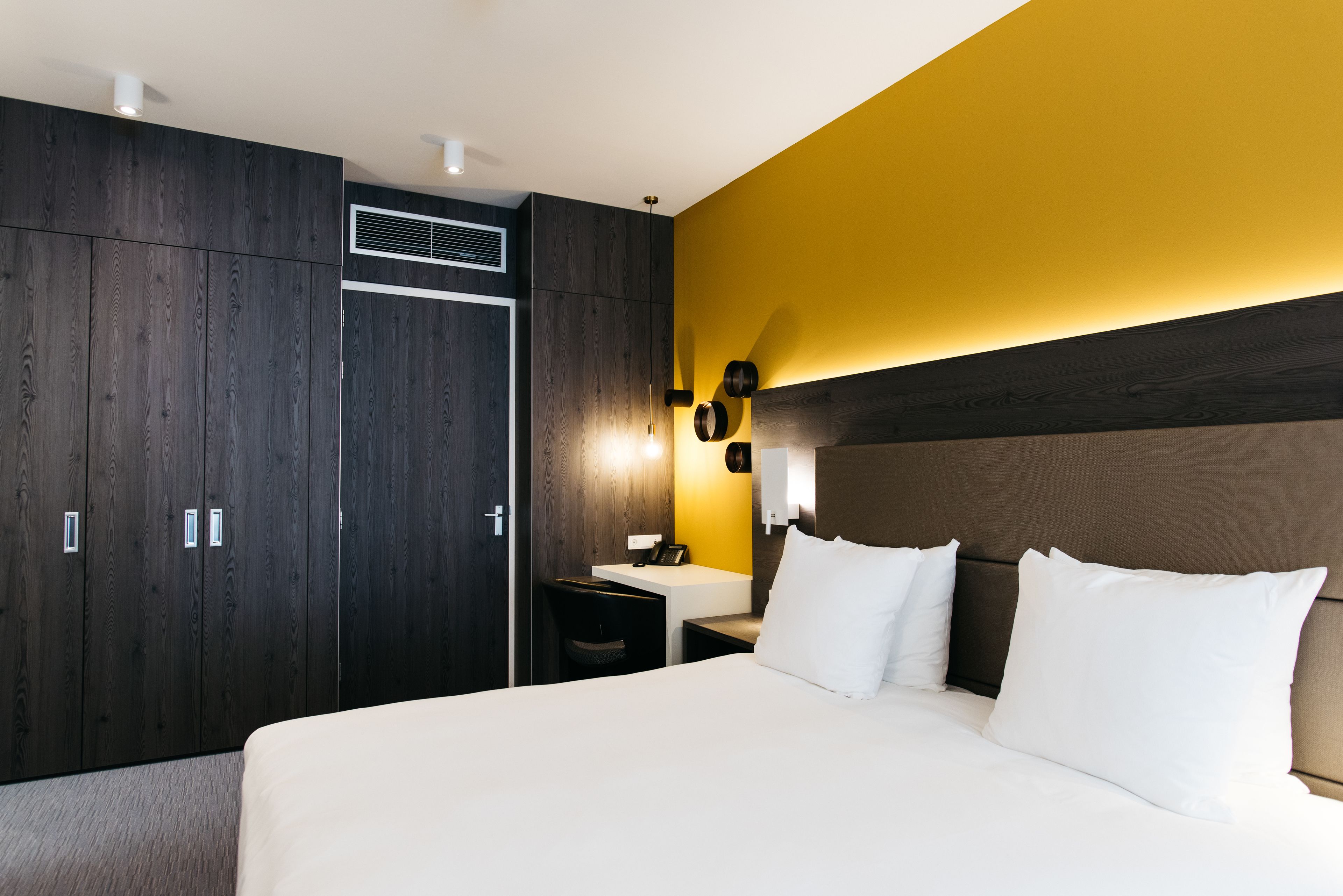 Habitació d'hotel amb un llit gran, una taula de treball amb cadira, un armari i una làmpada fixada a la paret.