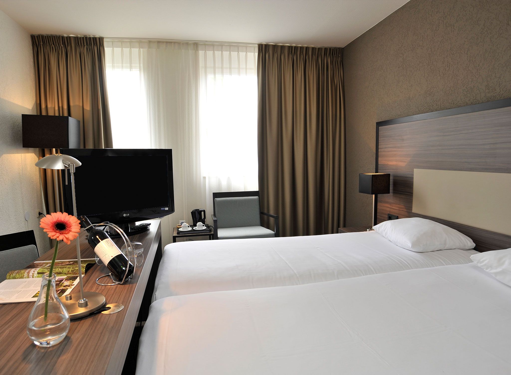 A hotel room with a bed, a desk with a TV, a chair, a nightstand, and a lamp.
