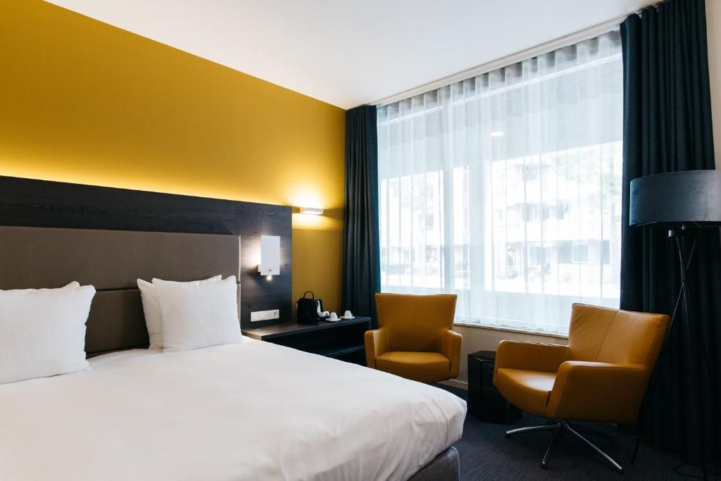 Een hotelkamer met een groot bed, twee fauteuils, een nachtkastje, een lamp en een raam met gordijnen.