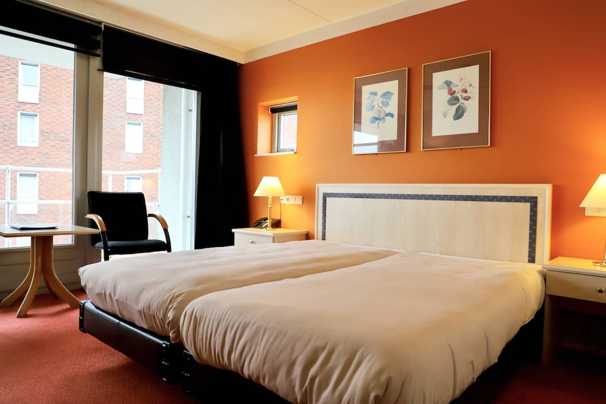 Ein Hotelzimmer mit einem großen Bett, einem Schreibtisch, einem Stuhl, zwei Nachttischen mit Lampen und einem Fenster mit Blick auf ein Backsteingebäude.