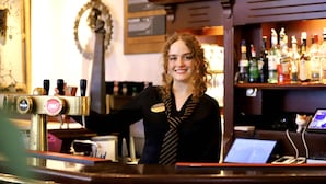 Un barista se tient devant une machine à café dans un bar, avec des étagères remplies de bouteilles et un ordinateur portable en arrière-plan.
