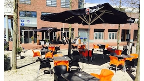Un espace extérieur aménagé avec des tables et des chaises, un espace couvert avec un parasol noir, et un bâtiment en arrière-plan.
