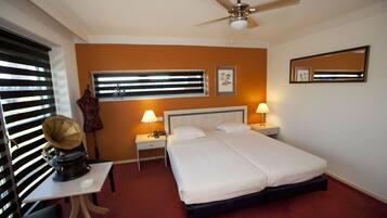 A hotel room with a bed, two bedside tables, a ceiling fan, a window with blinds, a desk with a lamp, and a mirror on the wall.