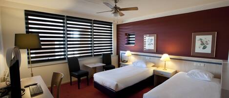 Ein Hotelzimmer mit zwei Betten, einem Schreibtisch mit Computer, einem Stuhl, einem kleinen Tisch und einem Deckenventilator.