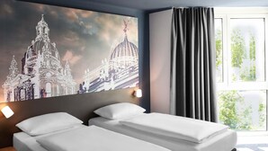 A hotel room with two beds, a large wall mural of a cathedral, a window with curtains, and a nightstand with a lamp. - B&B HOTEL Dresden City-Süd (Dresden)