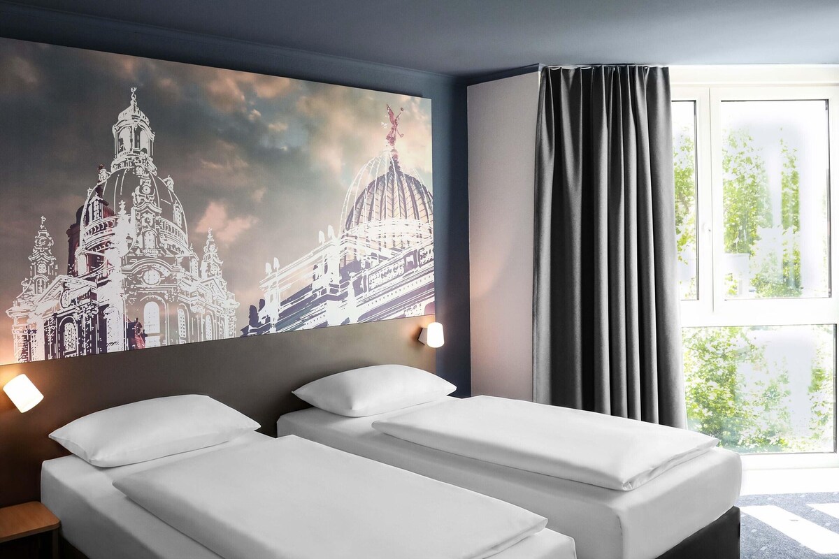 Ein Hotelzimmer mit zwei Betten, einem großen Wandgemälde einer Kathedrale, einem Fenster mit Vorhängen und einem Nachttisch mit Lampe.