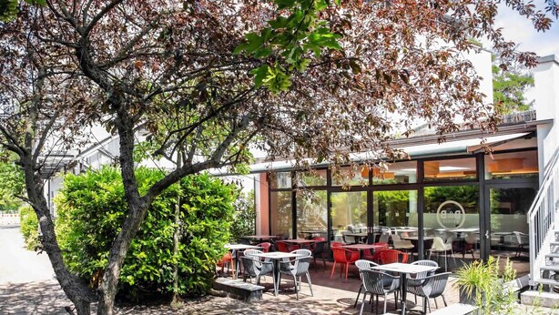 An outdoor patio area with tables and chairs, surrounded by greenery and a glass building. - B&B HOTEL Dresden City-Süd (Dresden)