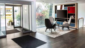 A modern interior with a glass door entrance, a seating area with chairs, a flat-screen TV, and a bookshelf. - B&B HOTEL Dresden City-Süd (Dresden)