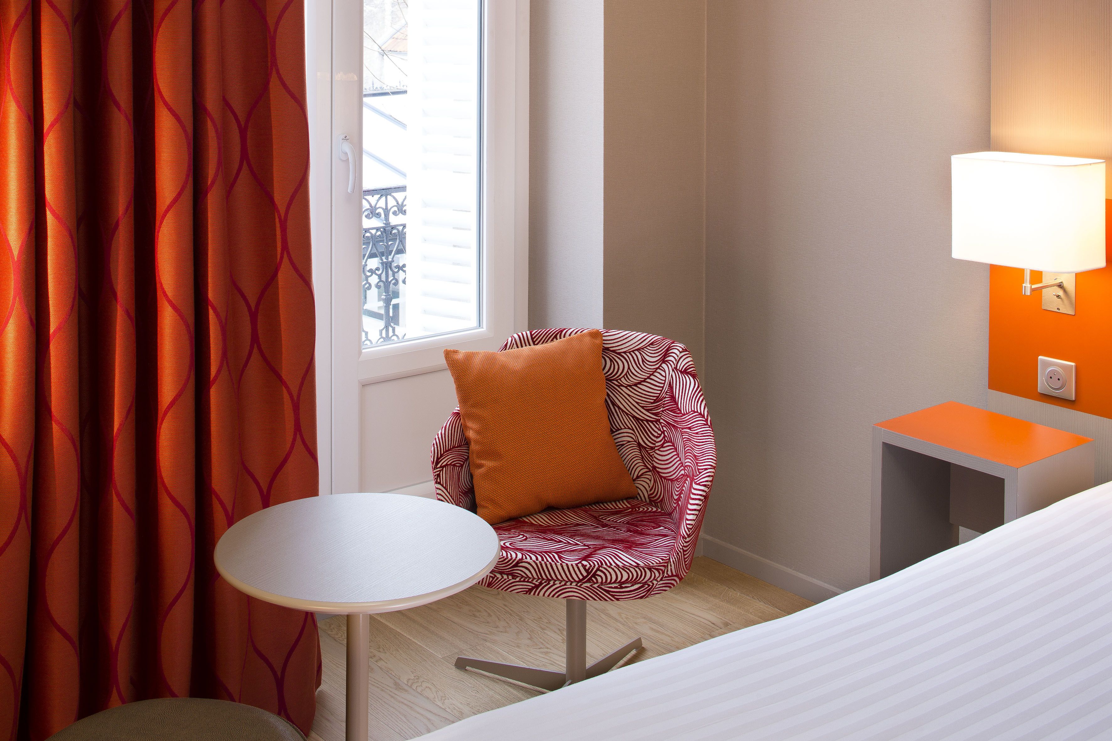 A hotel room with a bed, a small round table, a chair with an orange cushion, a window with curtains, and a wall-mounted lamp.